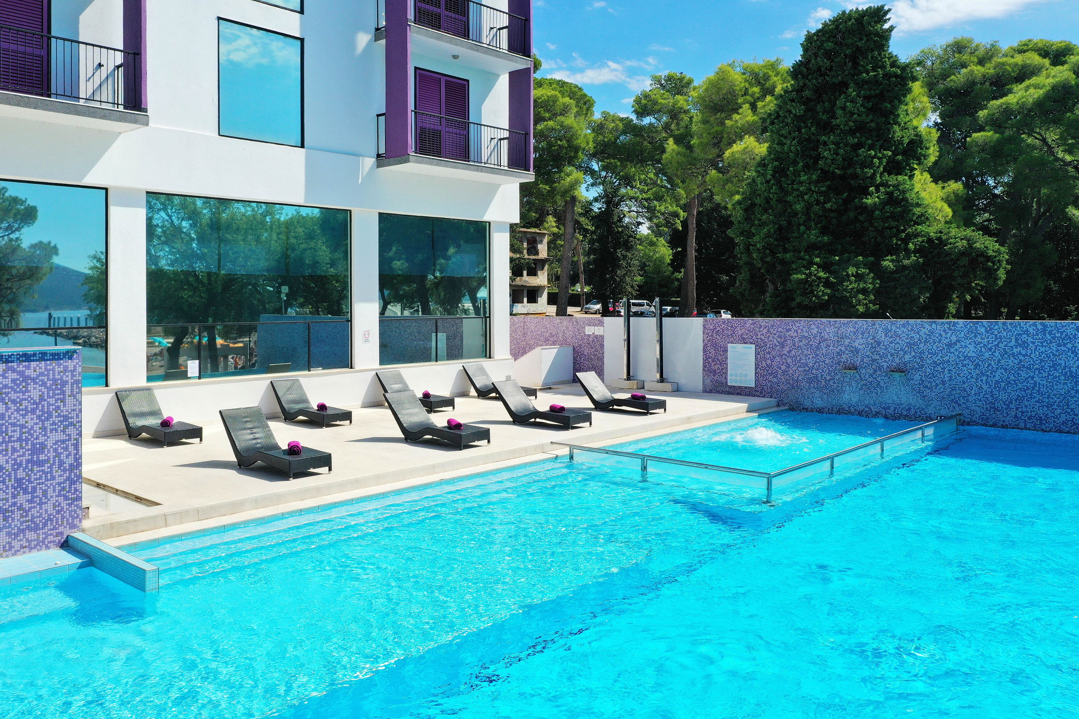 Une piscine avec des chaises longues et un bâtiment moderne doté de grandes fenêtres et d’éléments décoratifs violets.