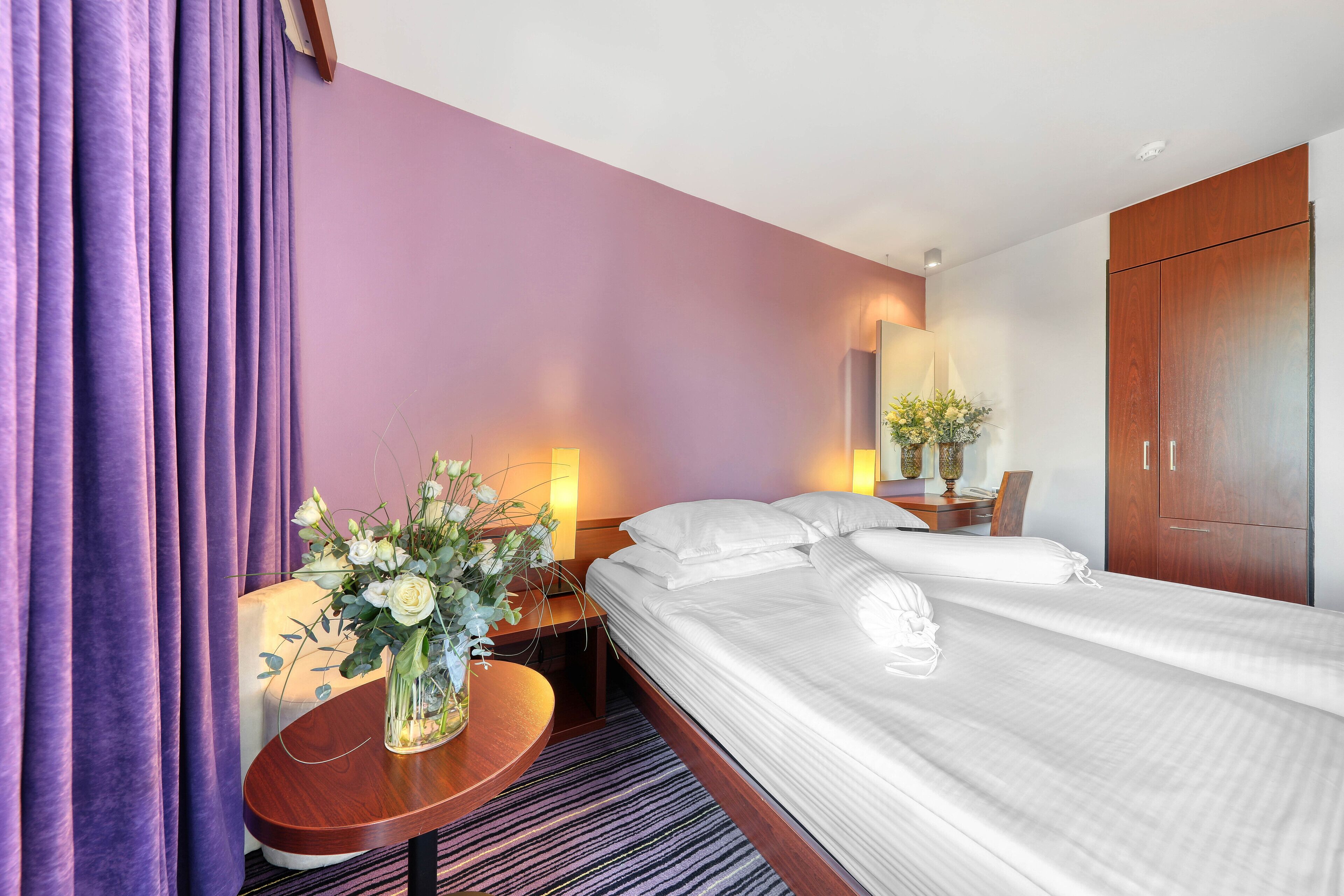 Ein Hotelzimmer mit zwei Betten, einem Holzschrank, einem runden Holztisch mit Blumenvase und einem Nachttisch mit Lampe.