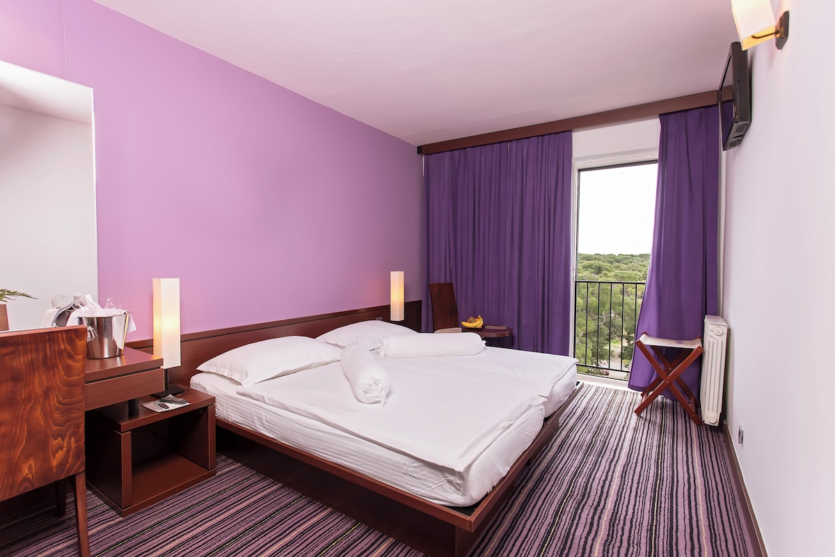 A hotel room with a large bed, a wooden desk, a nightstand, a lamp, a radiator, and purple curtains.