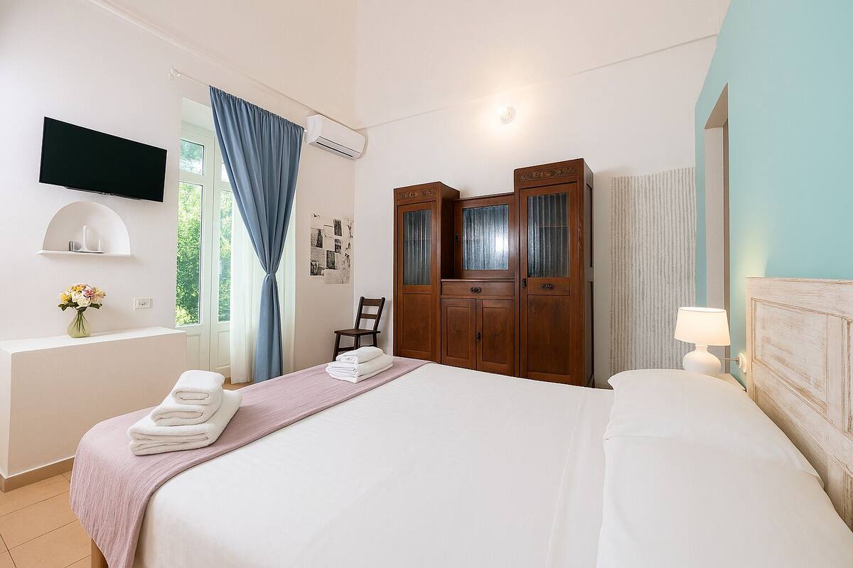 A hotel room with a bed, a desk with a chair, a vanity with a mirror, a TV mounted on the wall, and a window with curtains.