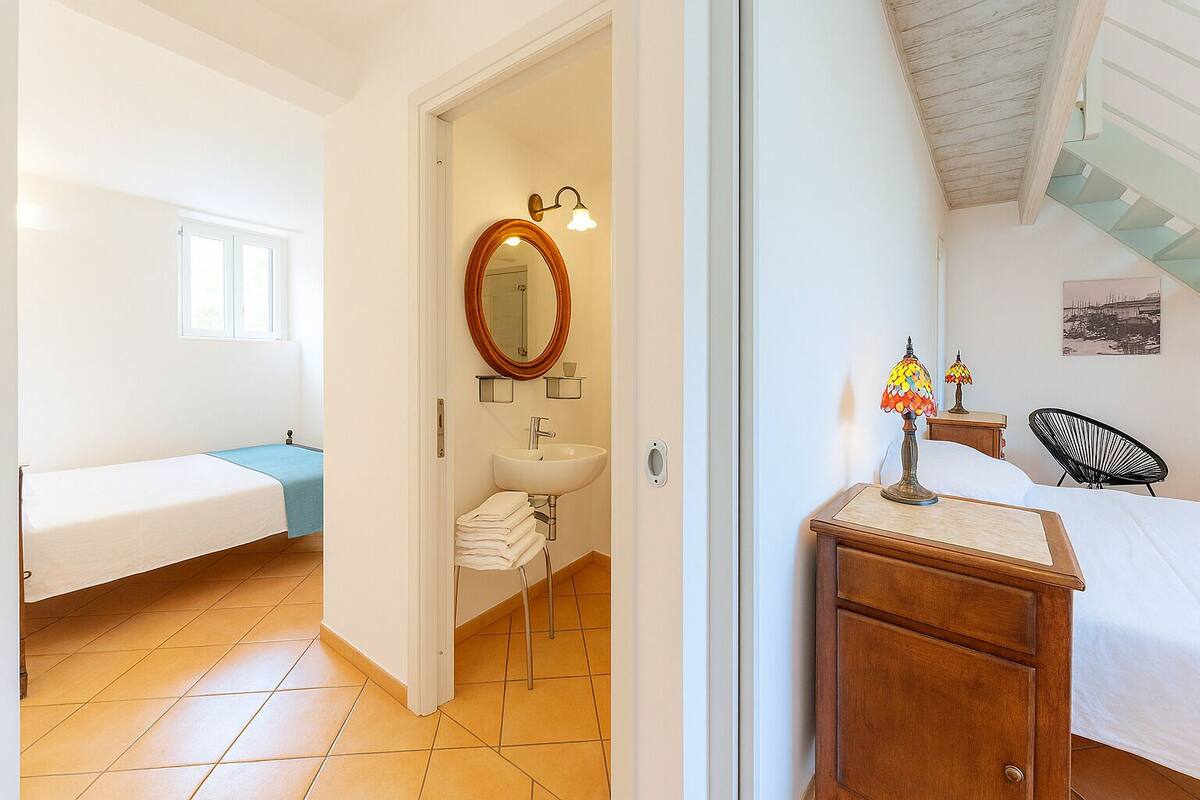 A bedroom with a bed, a bedside table with a lamp, a wooden dresser, and a bathroom with a sink and mirror.