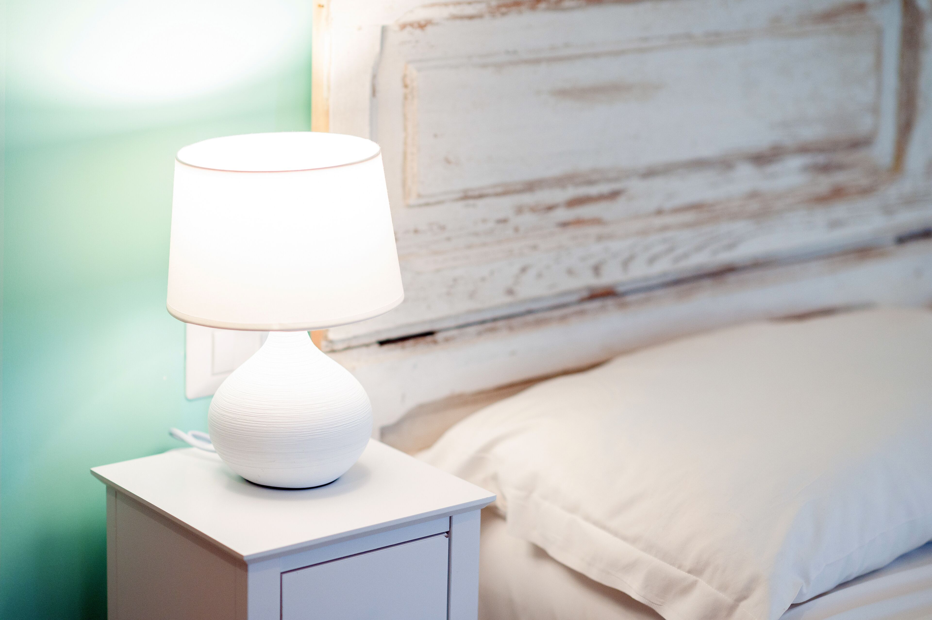 A bedside table with a white lamp, a bed with white bedding, and a green wall.