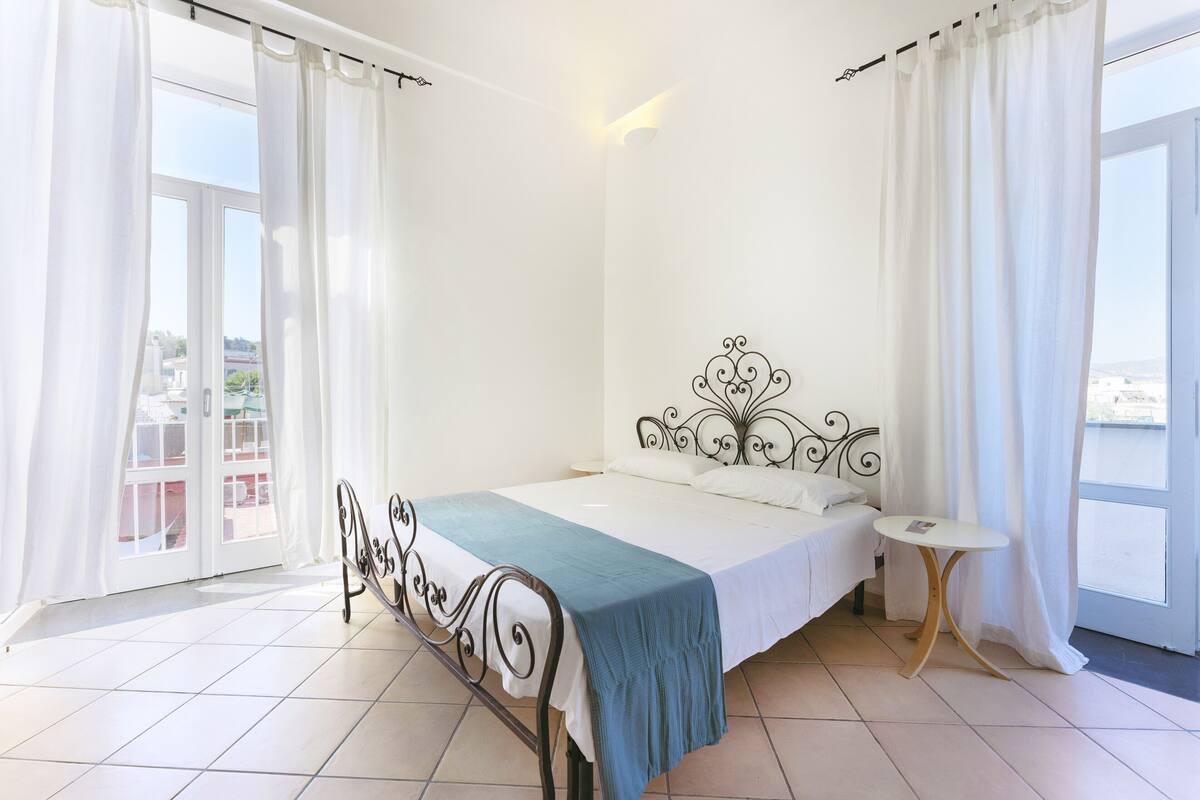 A bedroom with a metal bed frame, a bedside table, and a view through a large window.