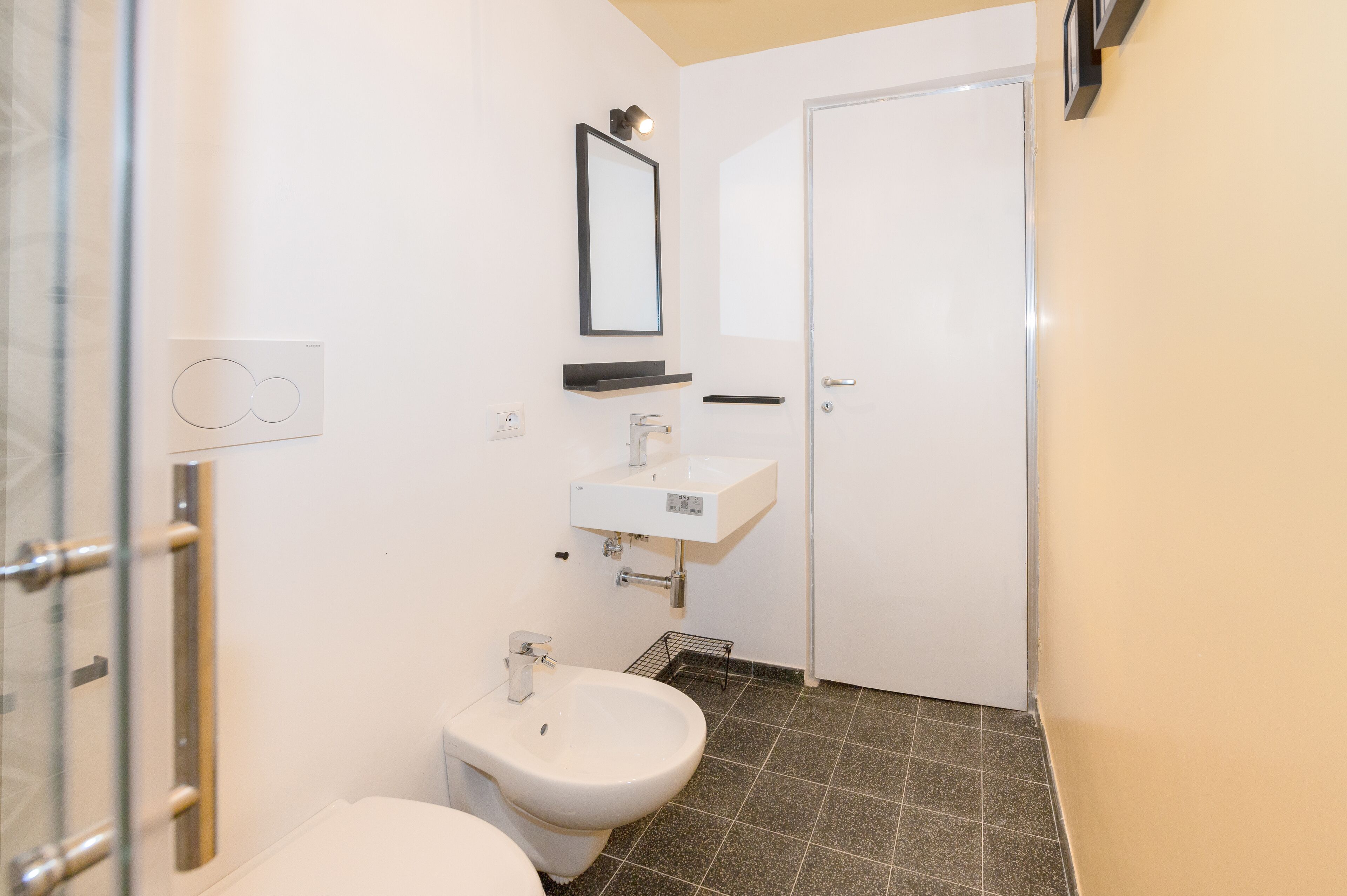 A bathroom with a toilet, bidet, sink, mirror, and a door.