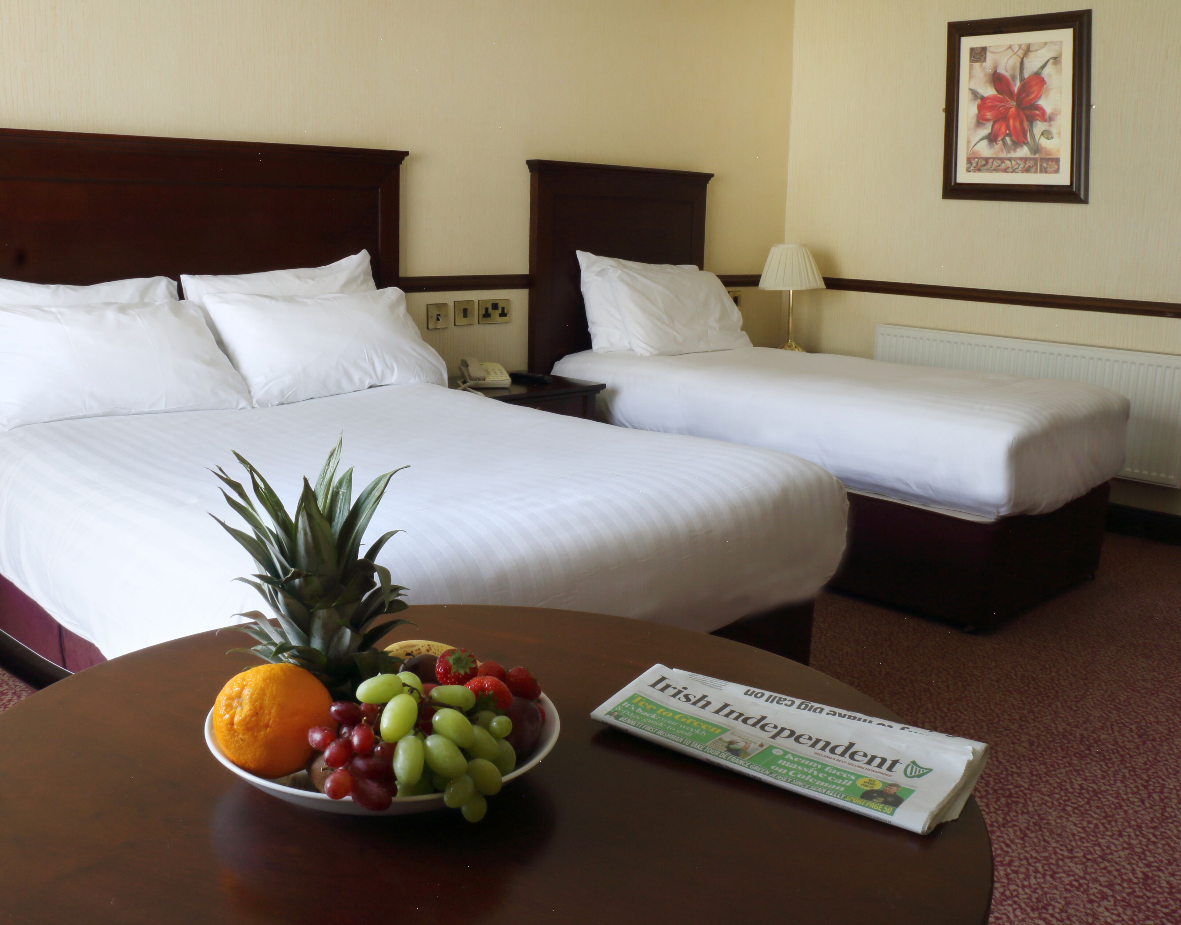Habitación de hotel con dos camas, una mesita de noche, una lámpara, un frutero y un periódico sobre la mesa.