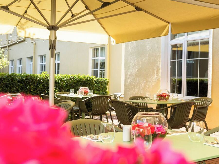 Een terras buiten met tafels en stoelen, een gele parasol en een gebouw op de achtergrond.
