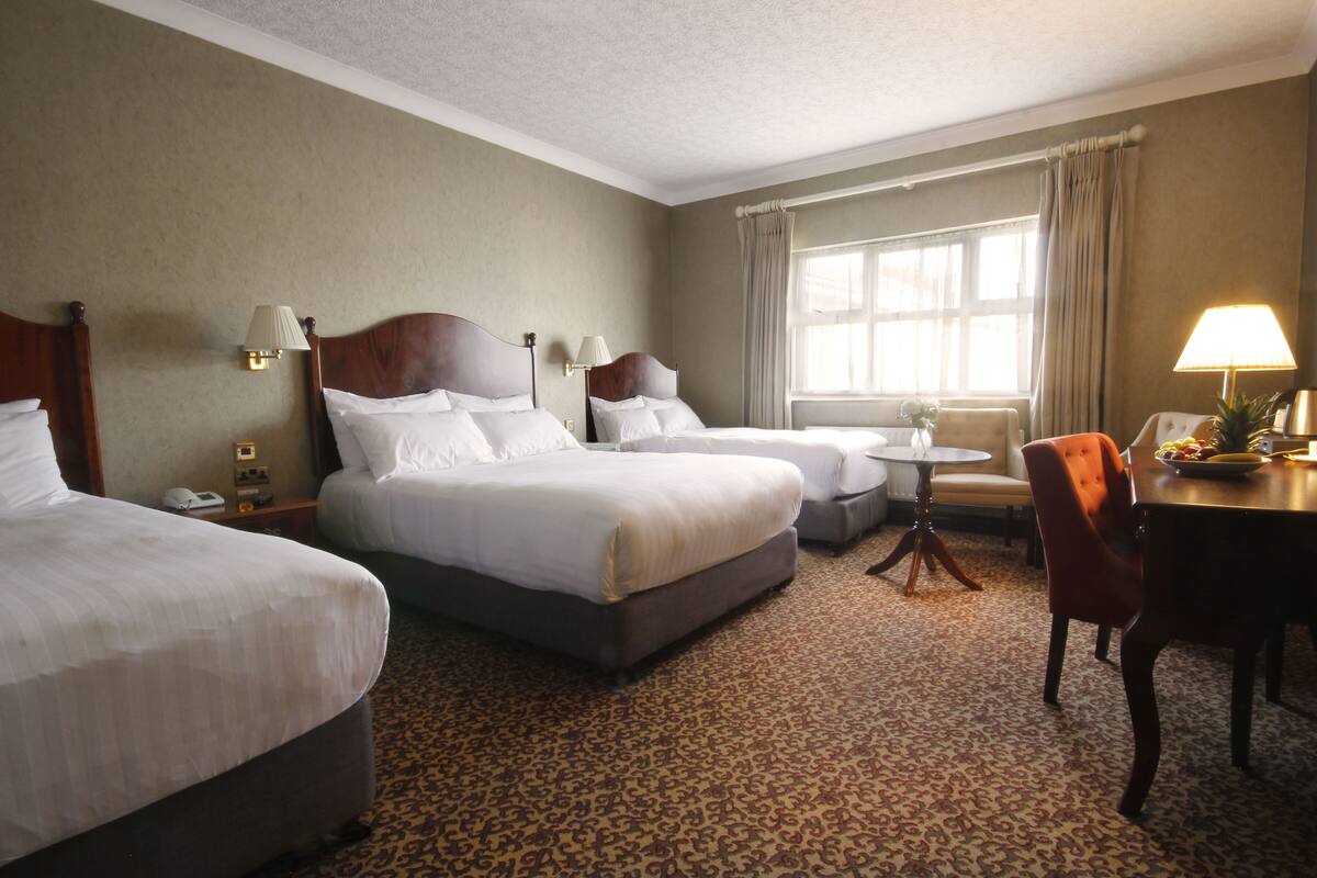 A hotel room with two beds, a small round table, a chair, and a window with curtains.