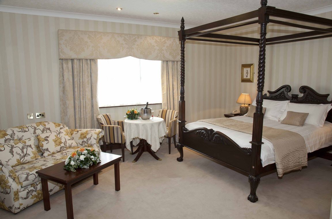 A four-poster bed with a canopy, a sofa, a round table with a vase of flowers, and a lamp on the bedside table.