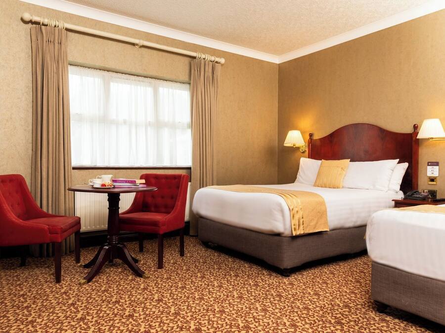 Habitación de hotel con dos camas, un sillón rojo, una mesita con libros y una ventana con cortinas.