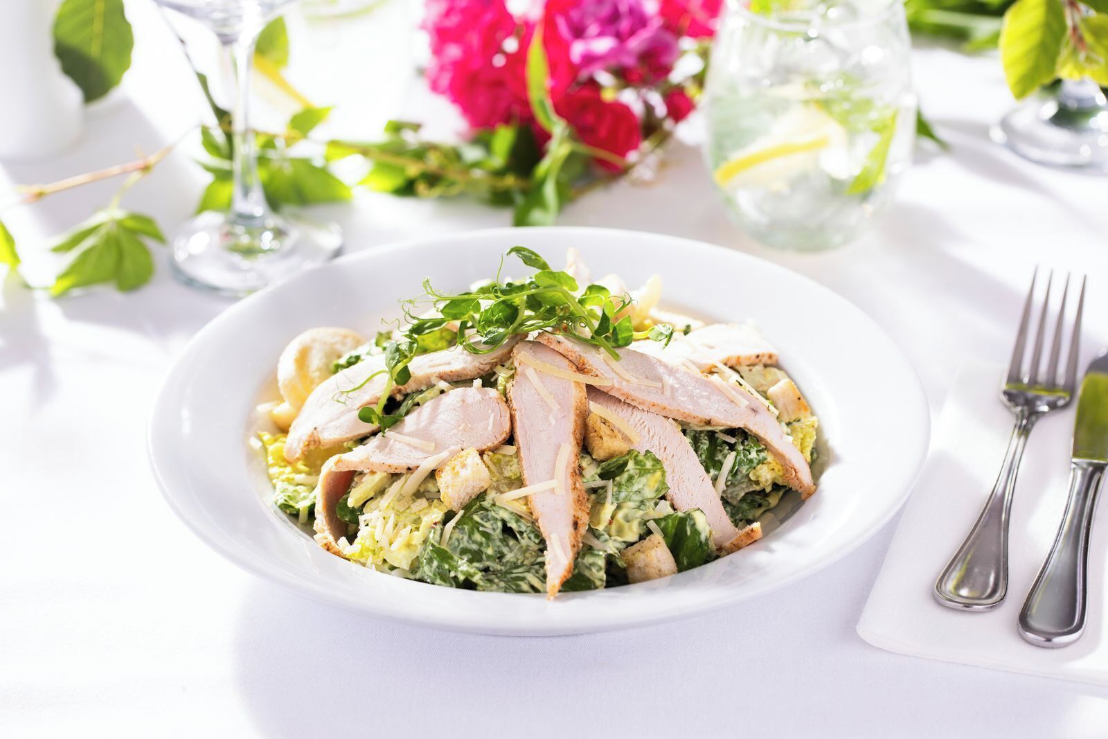 A plate of food with sliced meat and greens, accompanied by a fork and knife on a white tablecloth.