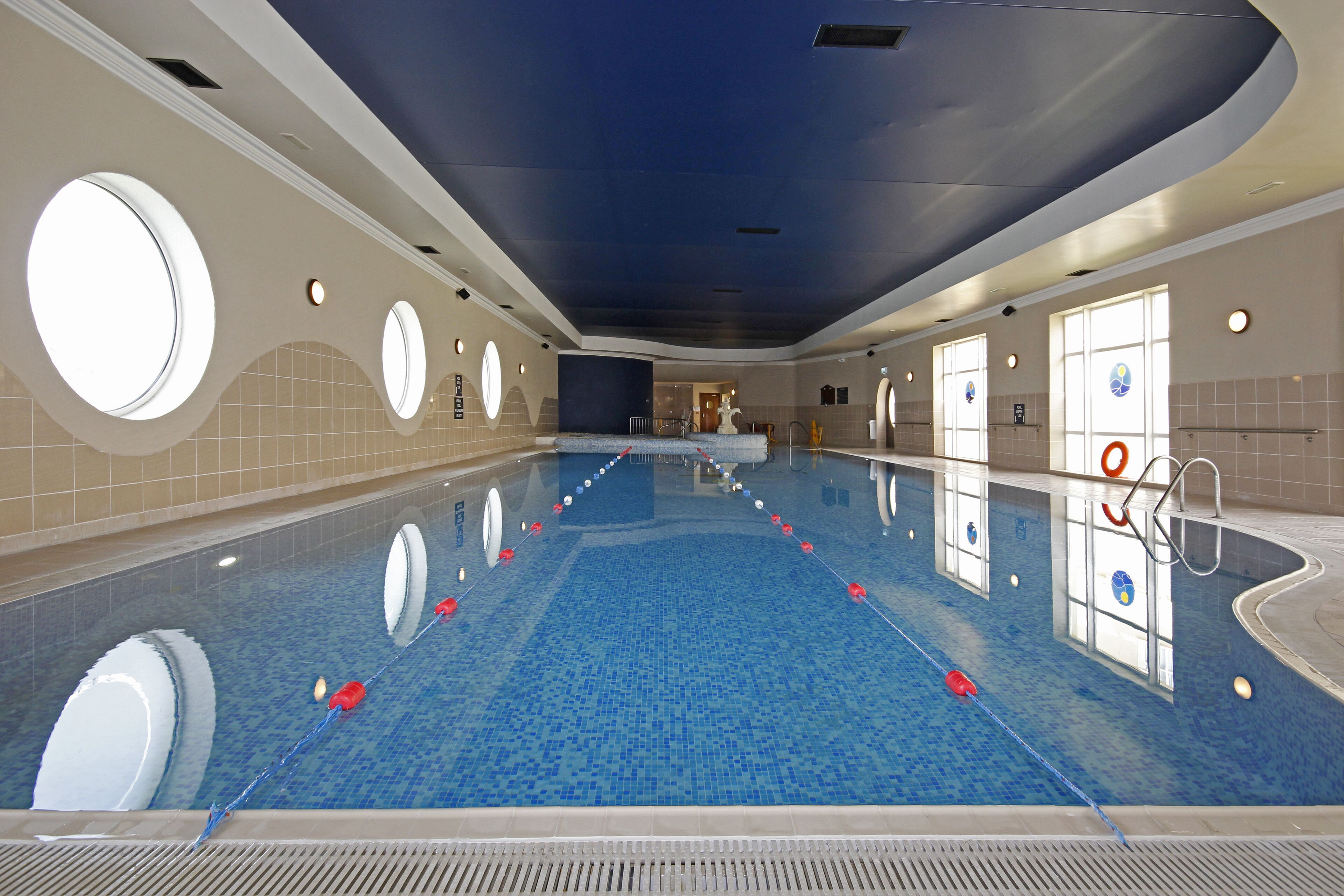A large indoor swimming pool with a blue tiled floor, circular windows, and a ceiling with recessed lighting.