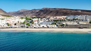 A coastal town with buildings along the beach, a clear view of the sea, and mountains in the background.