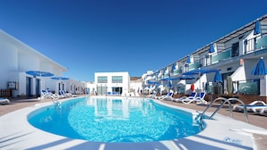 A pool area with sun loungers and umbrellas, a building with a white facade, and a clear blue sky. - Apartamentos Igramar MorroJable - Adults Only (Pajara)