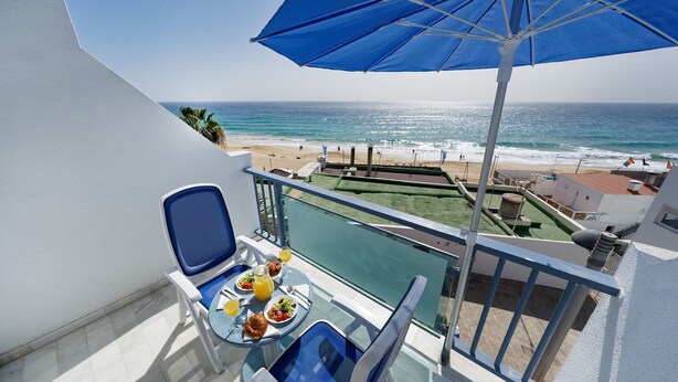 A balcony with a view of the beach and ocean, a table with breakfast, and a blue umbrella. - Apartamentos Igramar MorroJable - Adults Only (Pajara)