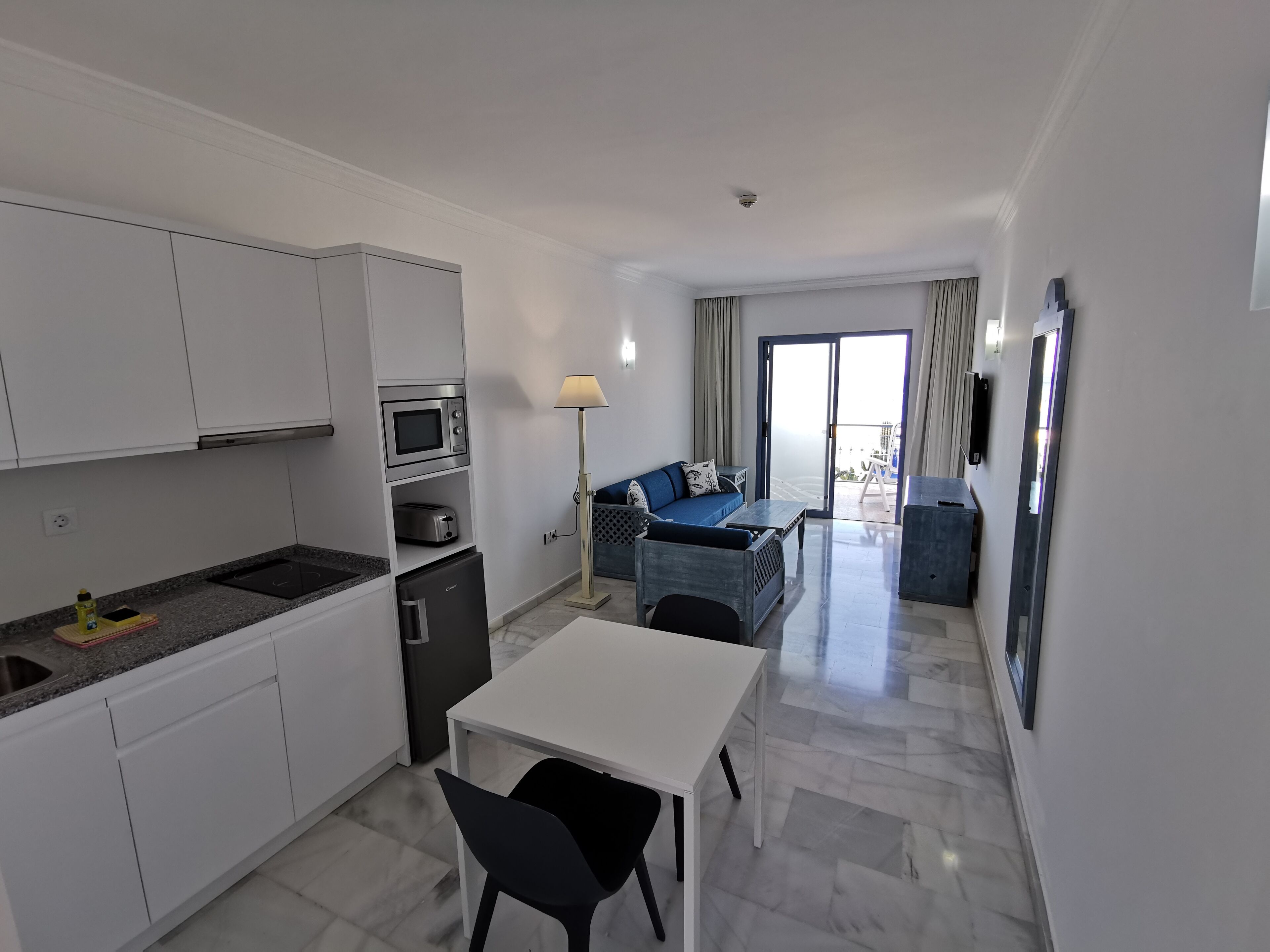 Cocina moderna con armarios blancos, un microondas y un refrigerador pequeño. Hay un comedor con una mesa blanca y sillas negras. En la sala de estar hay un sofá azul y una lámpara, con vistas al exterior a través de una puerta corrediza de vidrio.