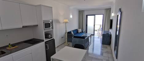 Cocina moderna con armarios blancos, un microondas y un refrigerador pequeño. Hay un comedor con una mesa blanca y sillas negras. En la sala de estar hay un sofá azul y una lámpara, con vistas al exterior a través de una puerta corrediza de vidrio.
