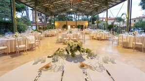 Ruim buitenterras met meerdere ronde tafels voor een formeel evenement, omringd door een overdekte paviljoen met betegelde vloer en uitzicht op de omliggende gebouwen en bomen.