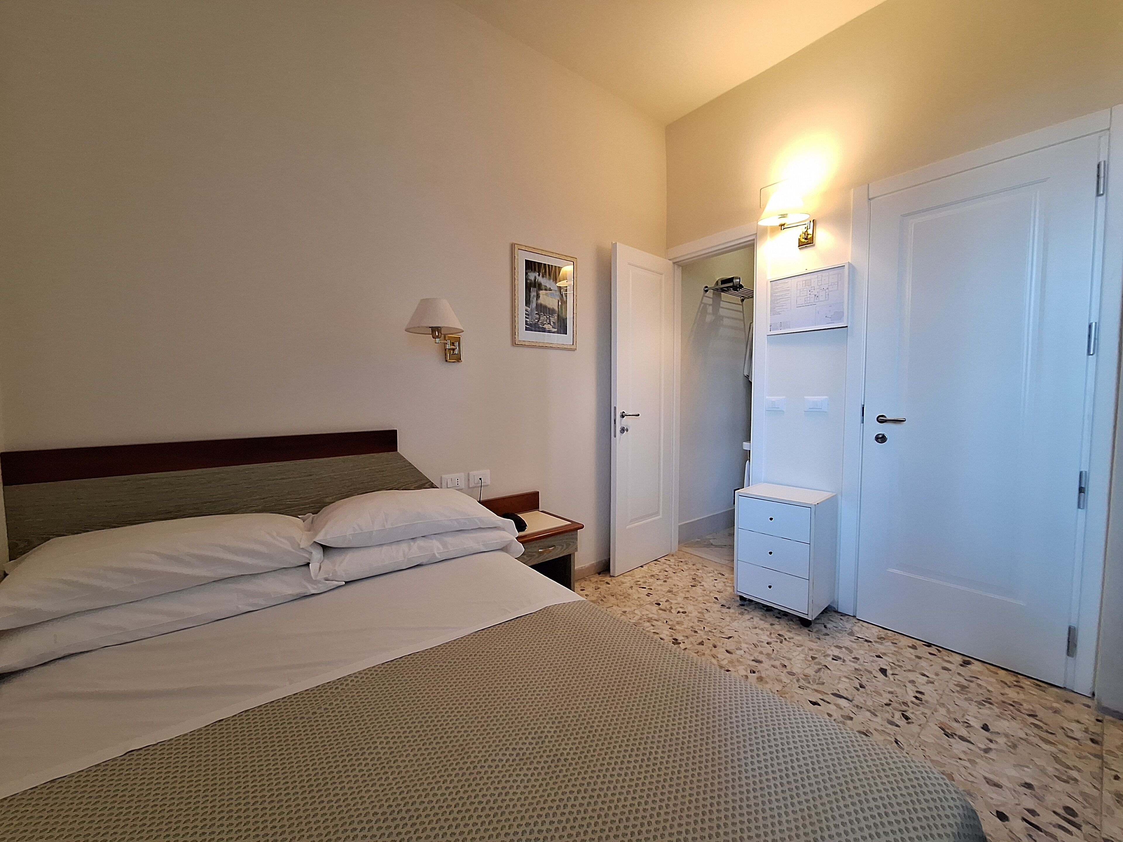 A hotel room with a bed, a nightstand, a lamp, a door, and a wall-mounted shelf.