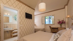 Een slaapkamer met een bed, een bureau, een stoel, een televisie, een raam en een badkamer met een wastafel en spiegel.