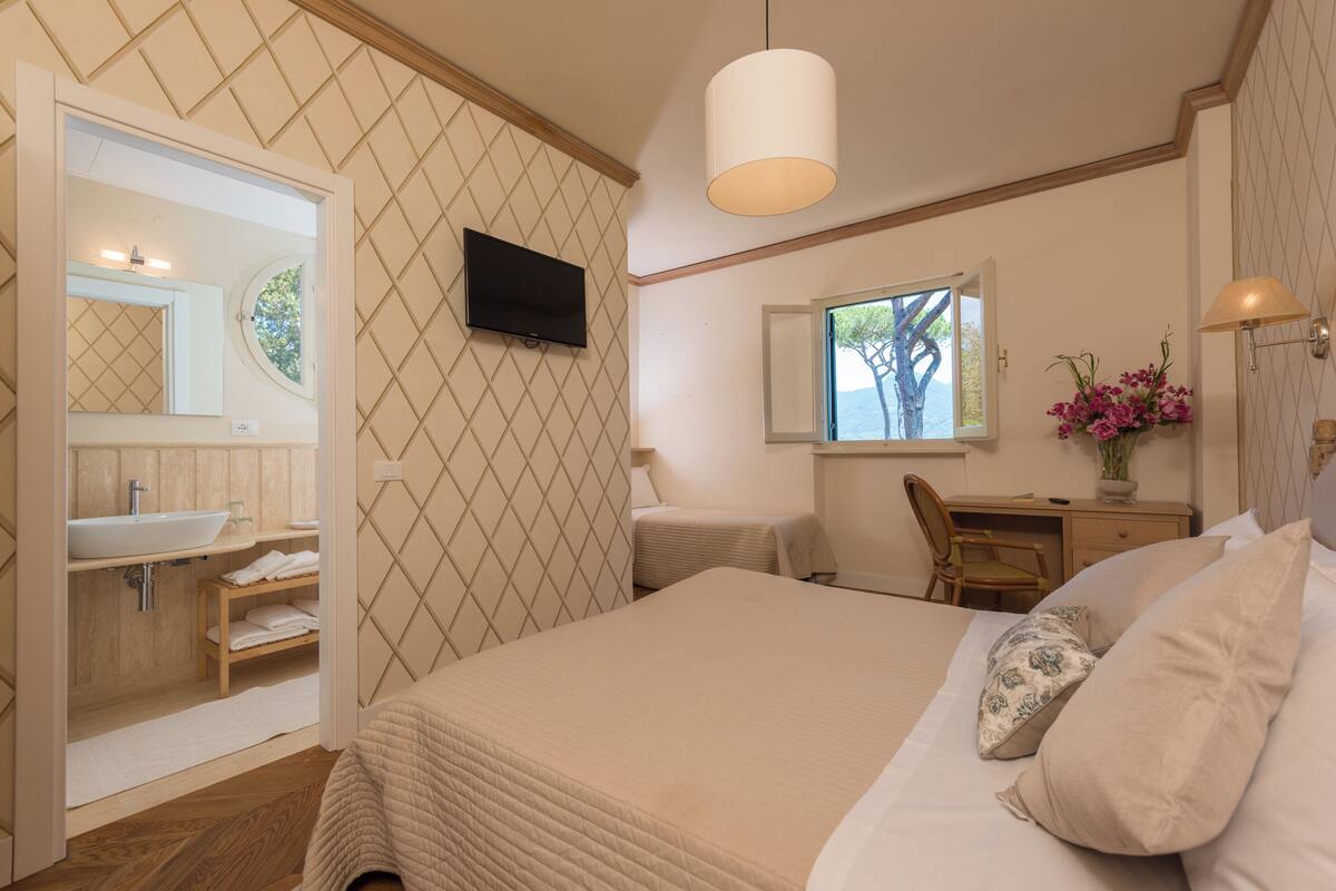 Een slaapkamer met een bed, een bureau, een stoel, een televisie, een raam en een badkamer met een wastafel en spiegel.