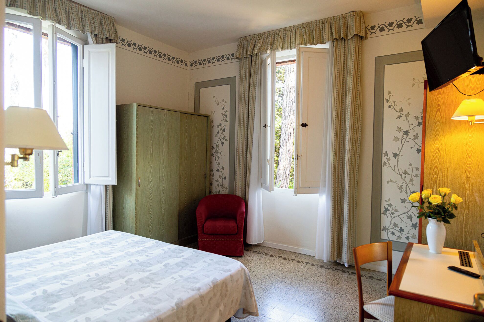 A bedroom with a bed, a red chair, a table with a vase of flowers, and a television.