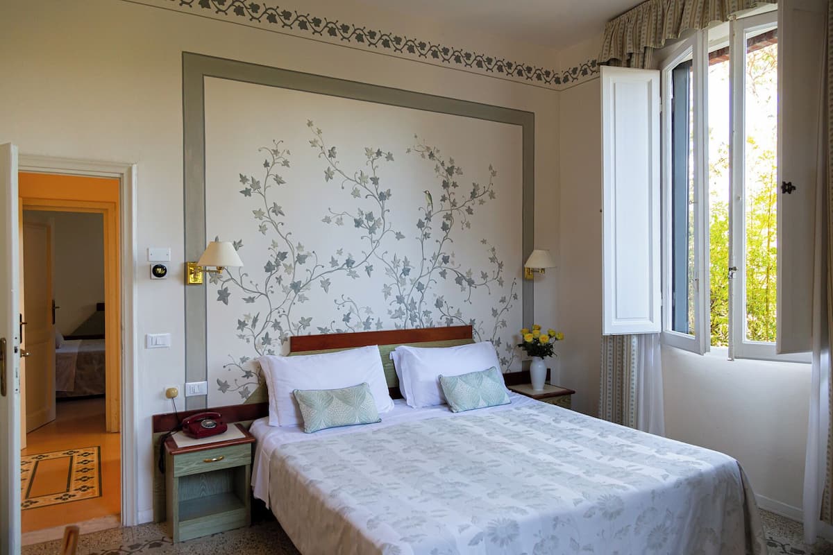 Een slaapkamer met een bed, nachtlampjes, een nachtkastje, een raam met gordijnen en een deur die naar een andere kamer leidt.