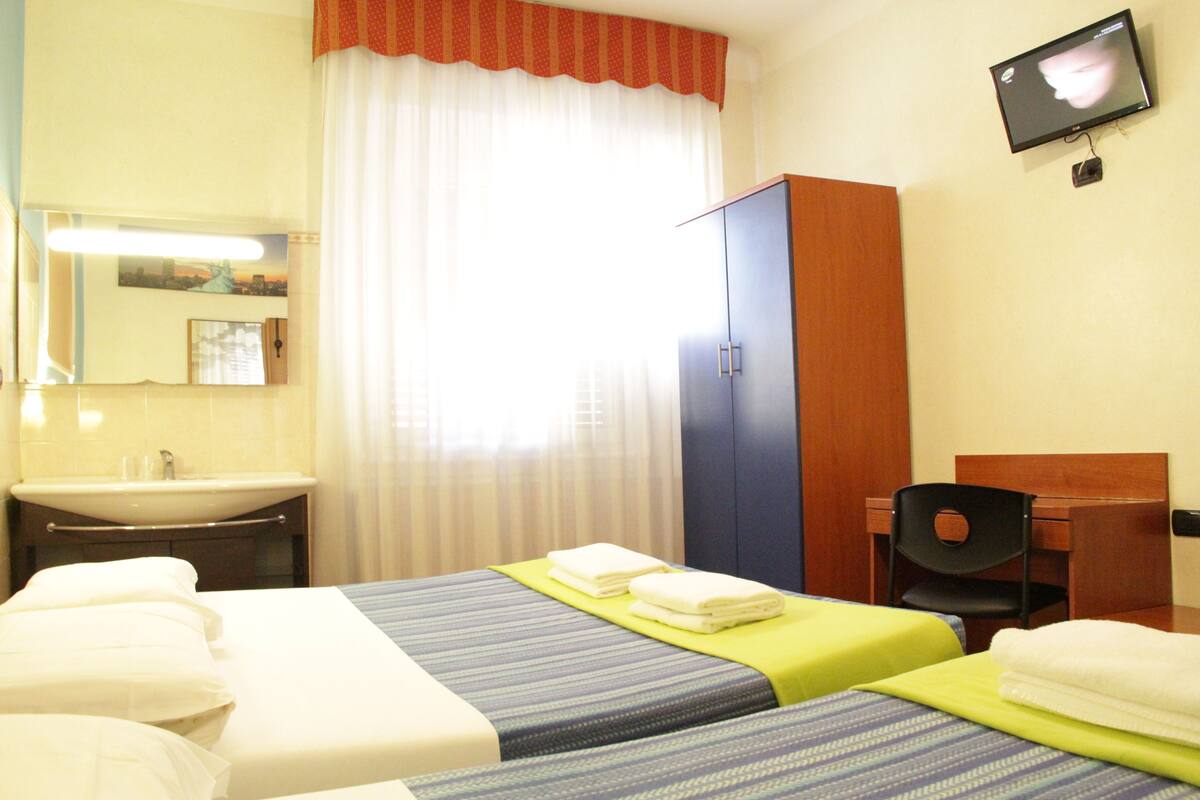 Camera d'albergo con due letti, una scrivania, una sedia, una televisione, un bagno con lavandino e specchio, e una finestra con tende.