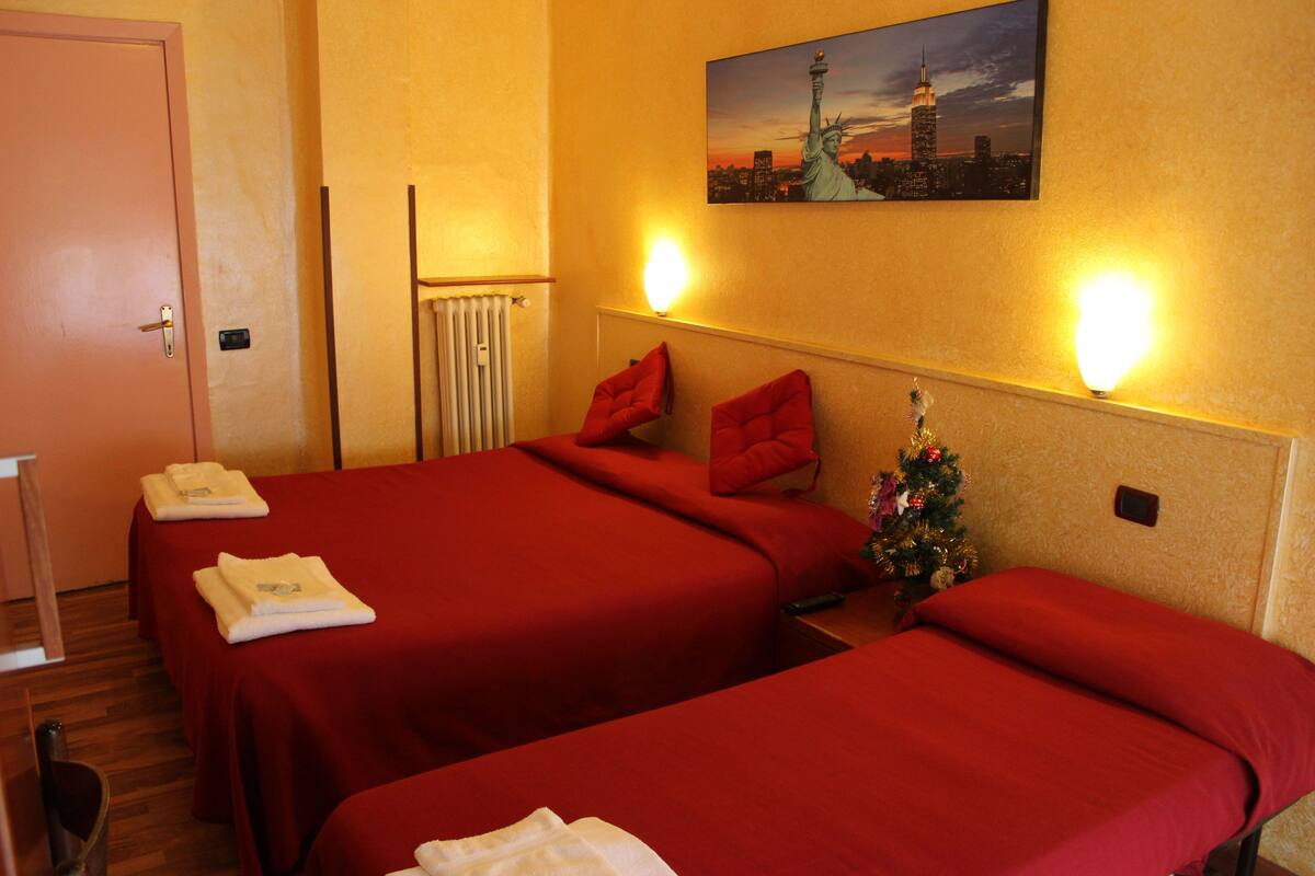 Camera d'albergo con due letti, biancheria rossa, un albero di Natale e un dipinto che raffigura un panorama urbano.