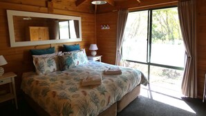 A wooden cabin bedroom with a bed, a nightstand, a lamp, a mirror, and a window with curtains. - Sandpiper Ocean Cottages (Douglas River)
