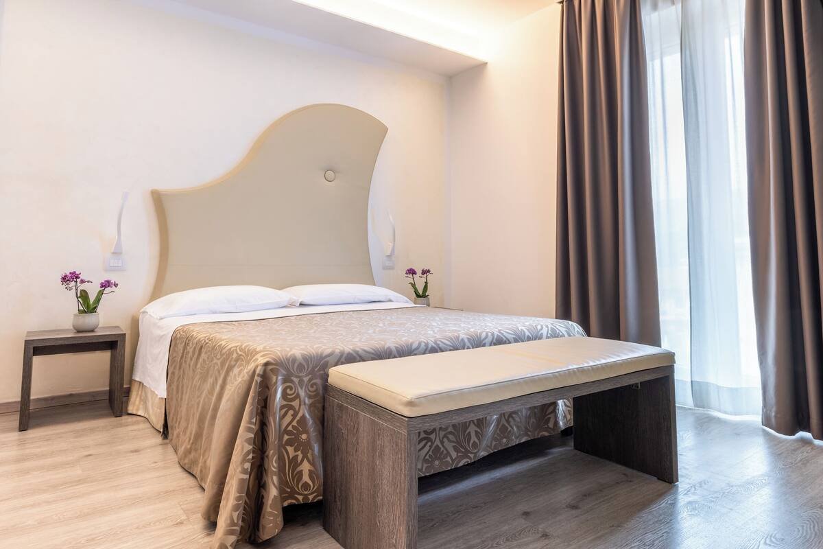 Habitación de hotel moderna con una cama grande, una mesita de noche con un jarrón de flores y un banco al pie de la cama.