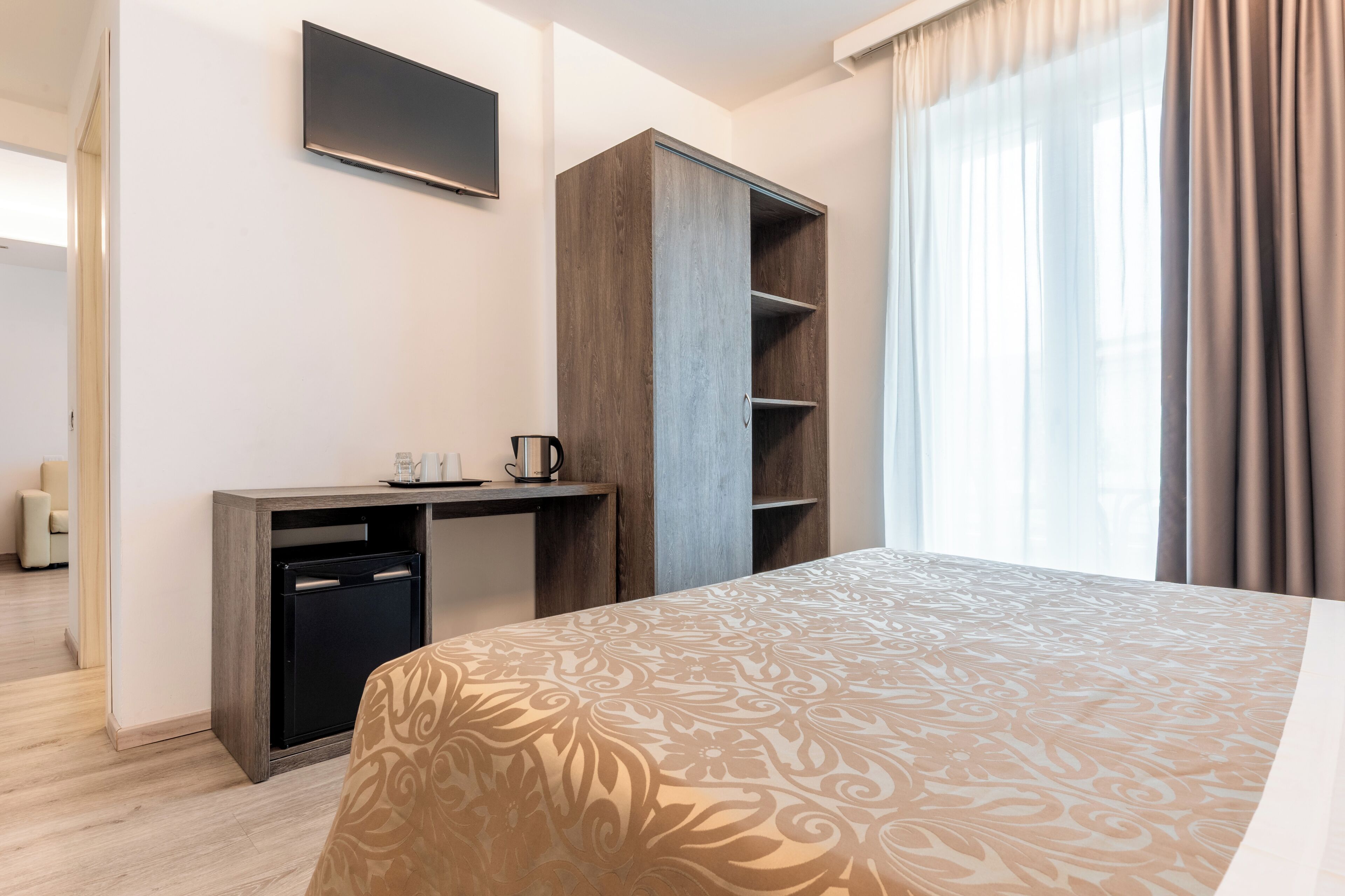 A hotel room with a bed, a desk with a coffee maker, a wardrobe, and a TV on the wall.