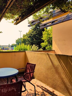Un patio con mesa y sillas, una pérgola arriba y vista a un estacionamiento y árboles.