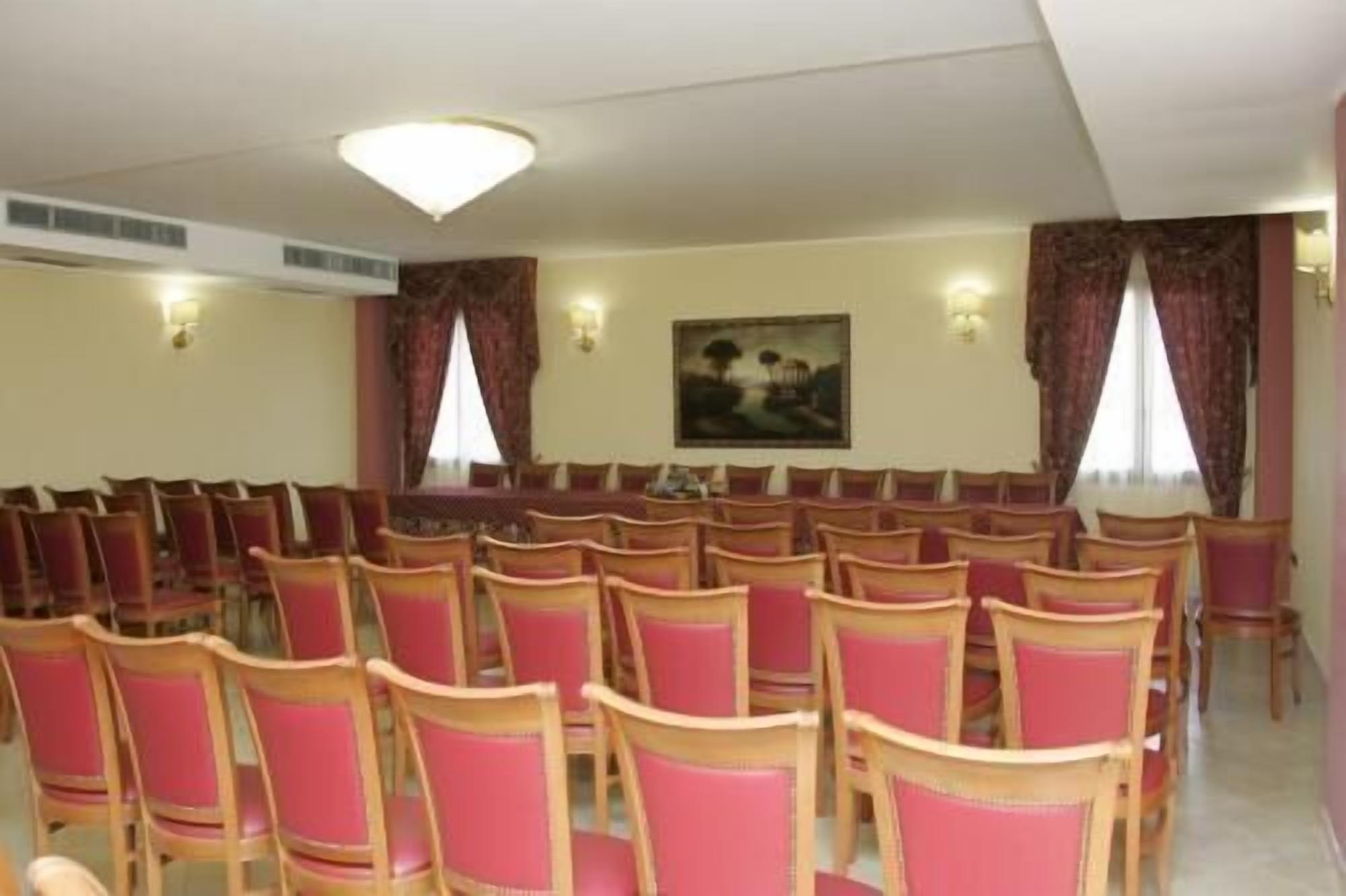 Una sala de conferències amb files de cadires de fusta i coixinets vermells, un quadre a la paret i dues finestres amb cortines.