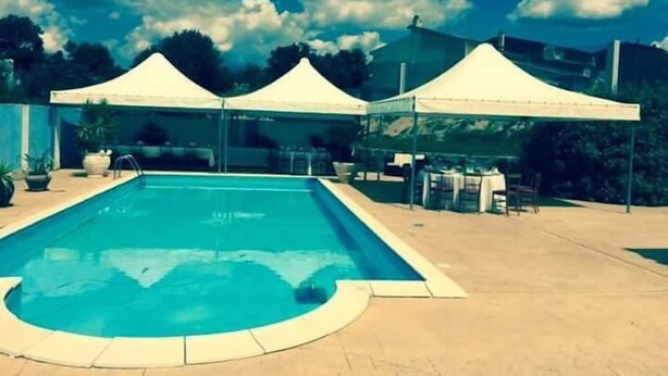 A swimming pool with a curved edge, surrounded by a tiled deck. There are two white tents in the background. - La Madonnina (Budduso)