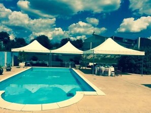 A swimming pool with a curved edge, surrounded by a tiled deck. There are two white tents in the background. - La Madonnina (Budduso)