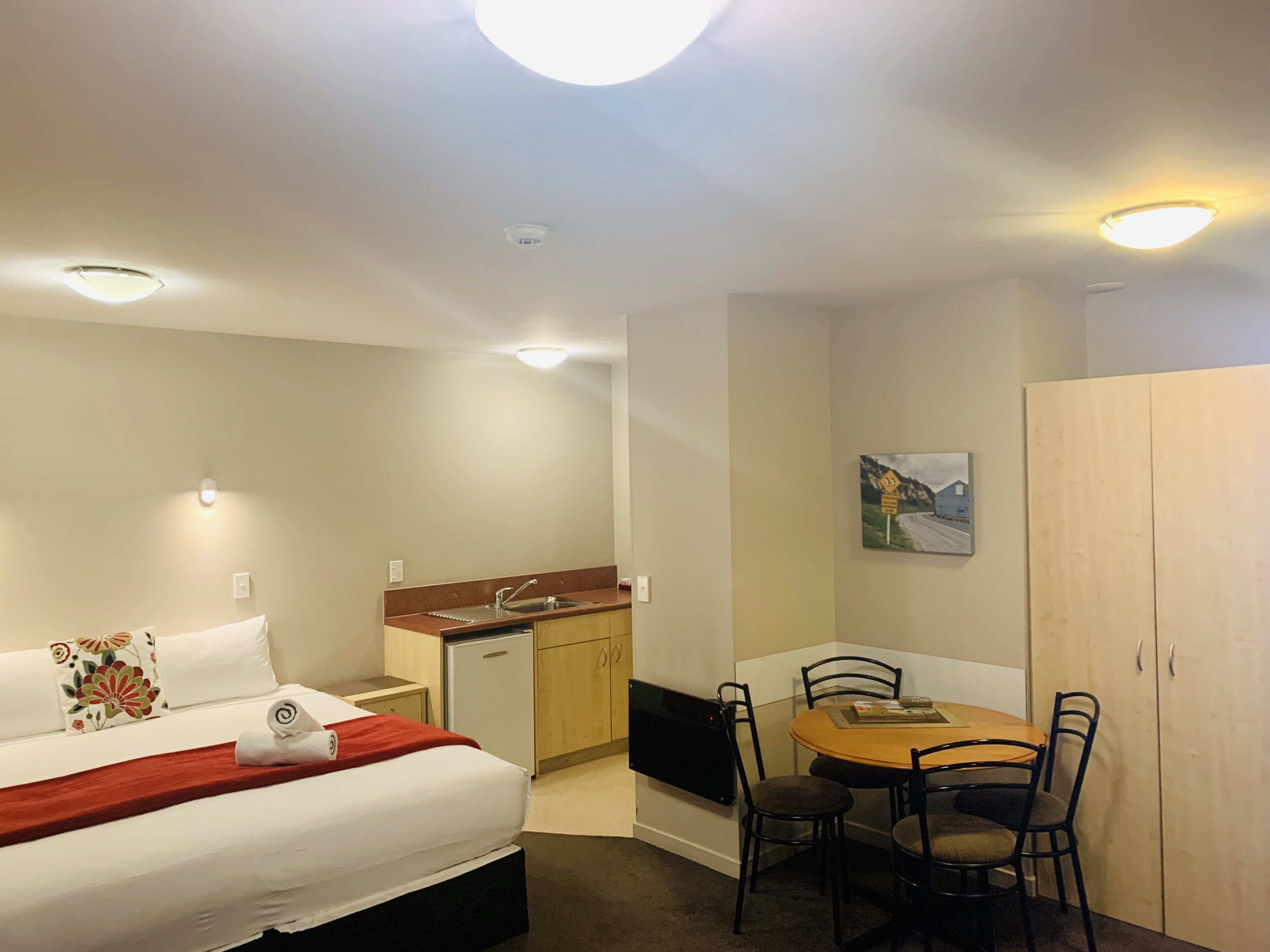 A hotel room with a bed, a small kitchenette, a dining table with chairs, and a painting on the wall.