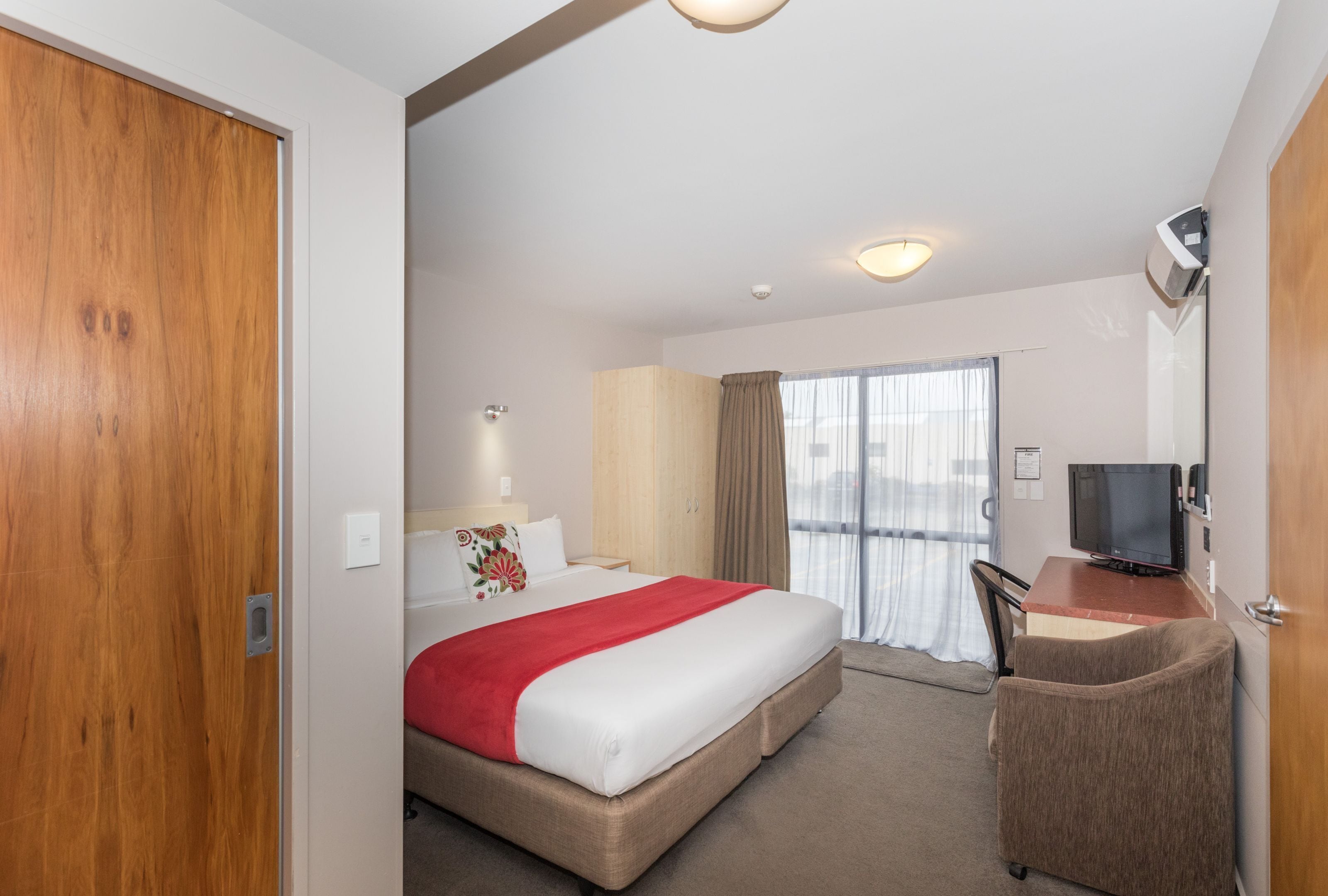 A hotel room with a bed, a desk with a TV, a chair, a window with curtains, and a door.
