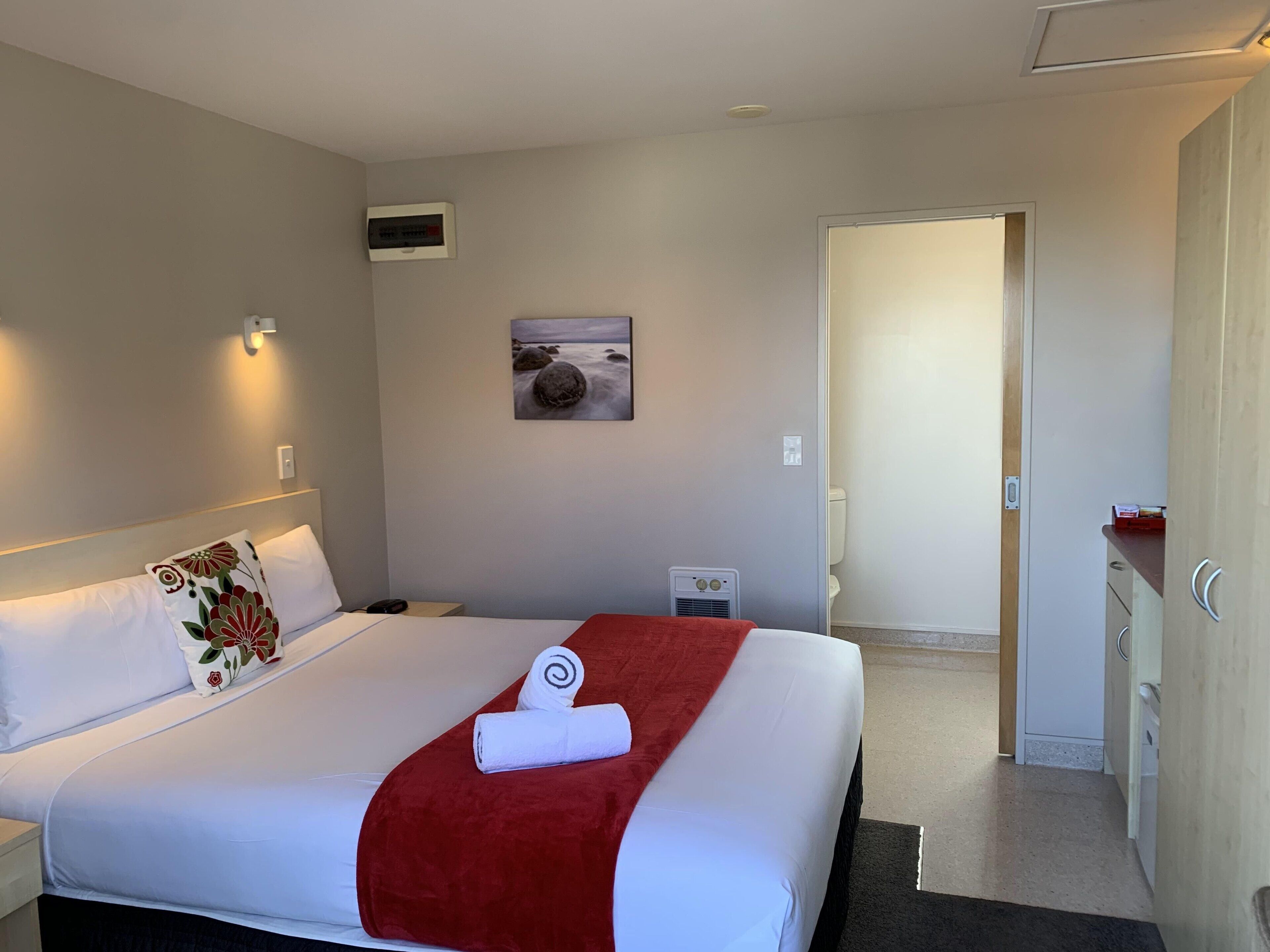 Habitación de hotel con una cama bien hecha, una mesita de noche pequeña, aire acondicionado instalado en la pared, un cuadro con una escena de playa y una puerta que da a un baño.