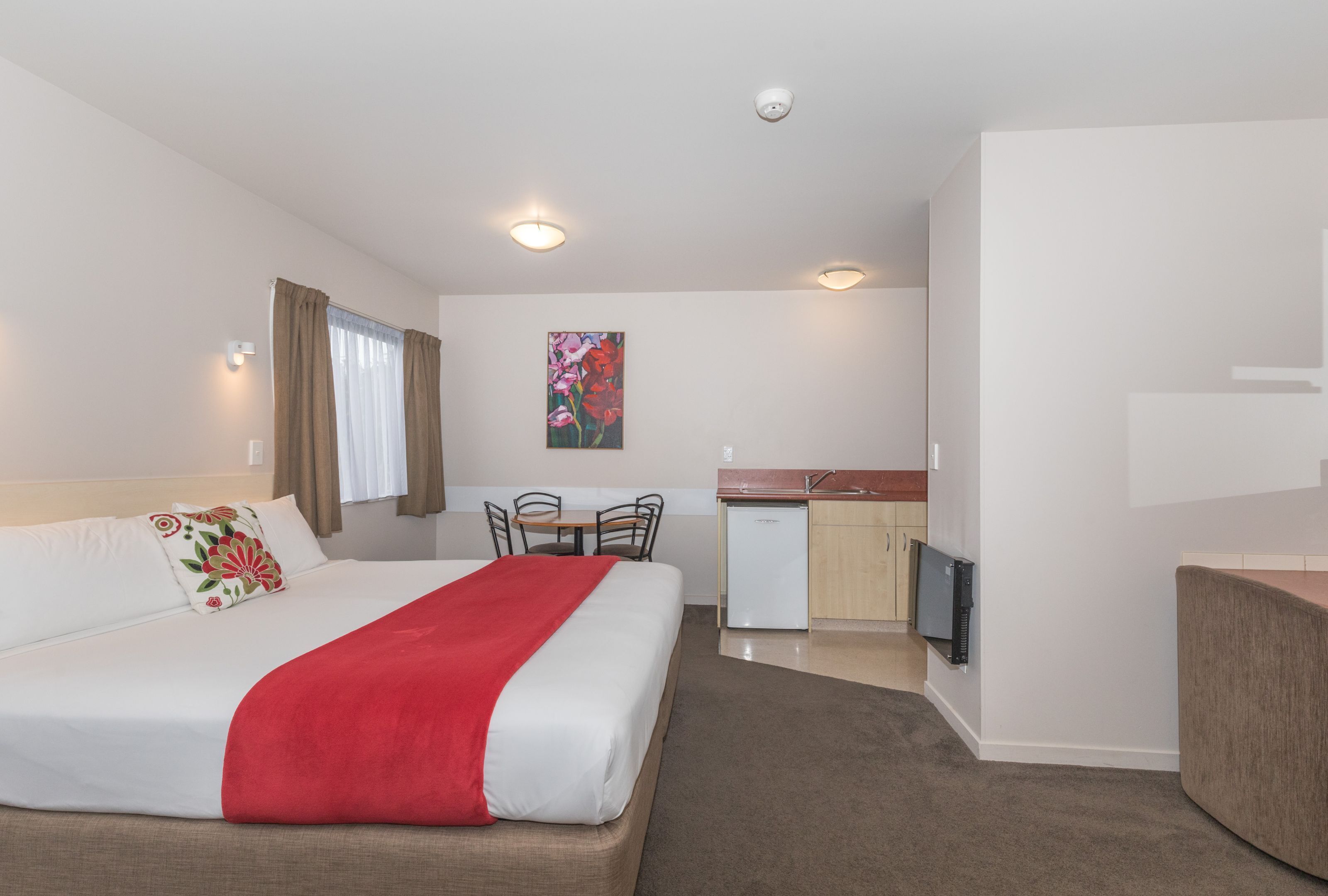 A hotel room with a bed, a desk, a chair, a small kitchenette, and a TV.