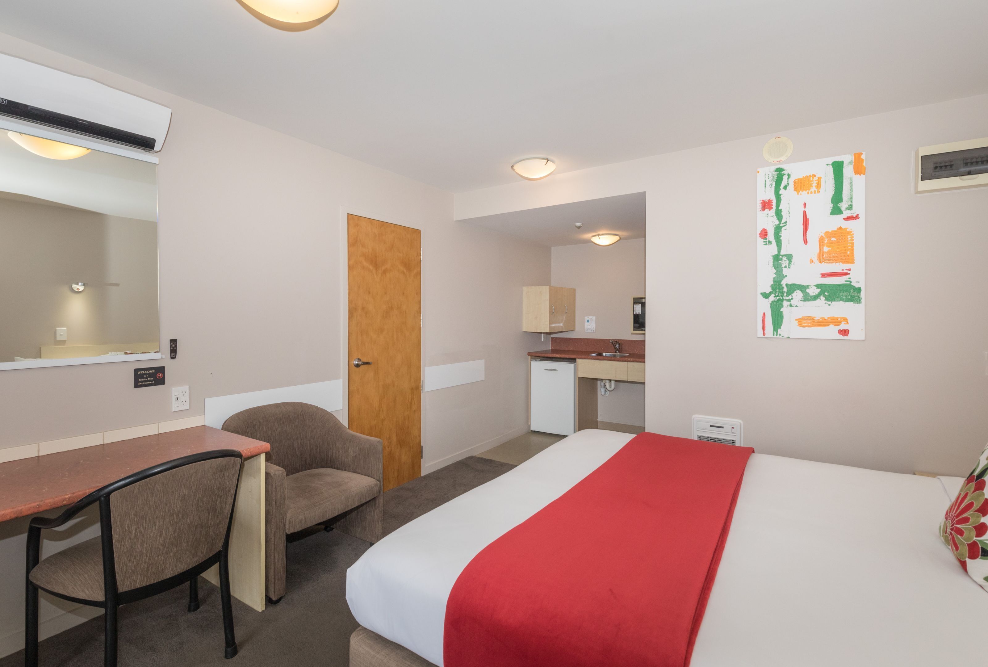A hotel room with a bed, a desk, a chair, a small kitchenette, and a bathroom.