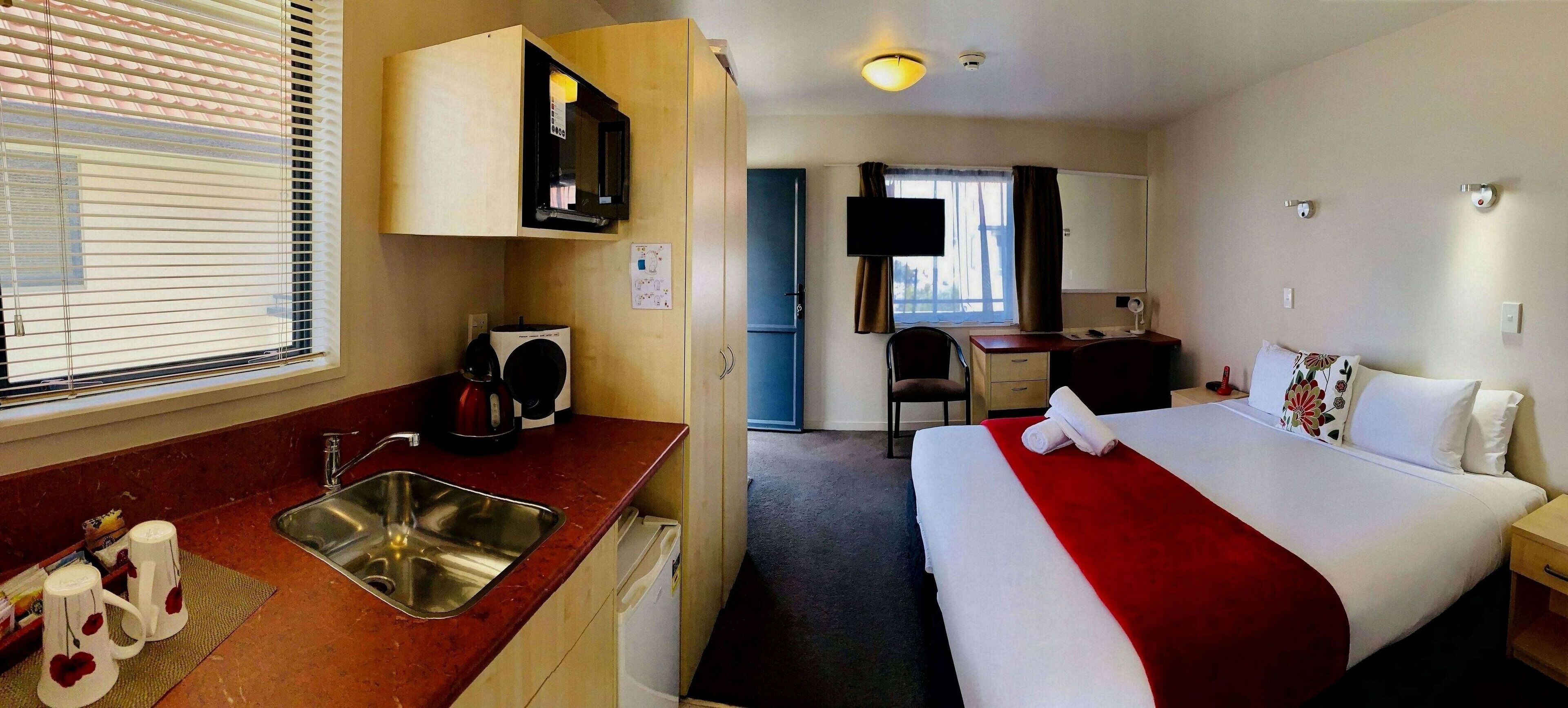 A hotel room with a bed, a desk, a chair, a TV, and a kitchenette.