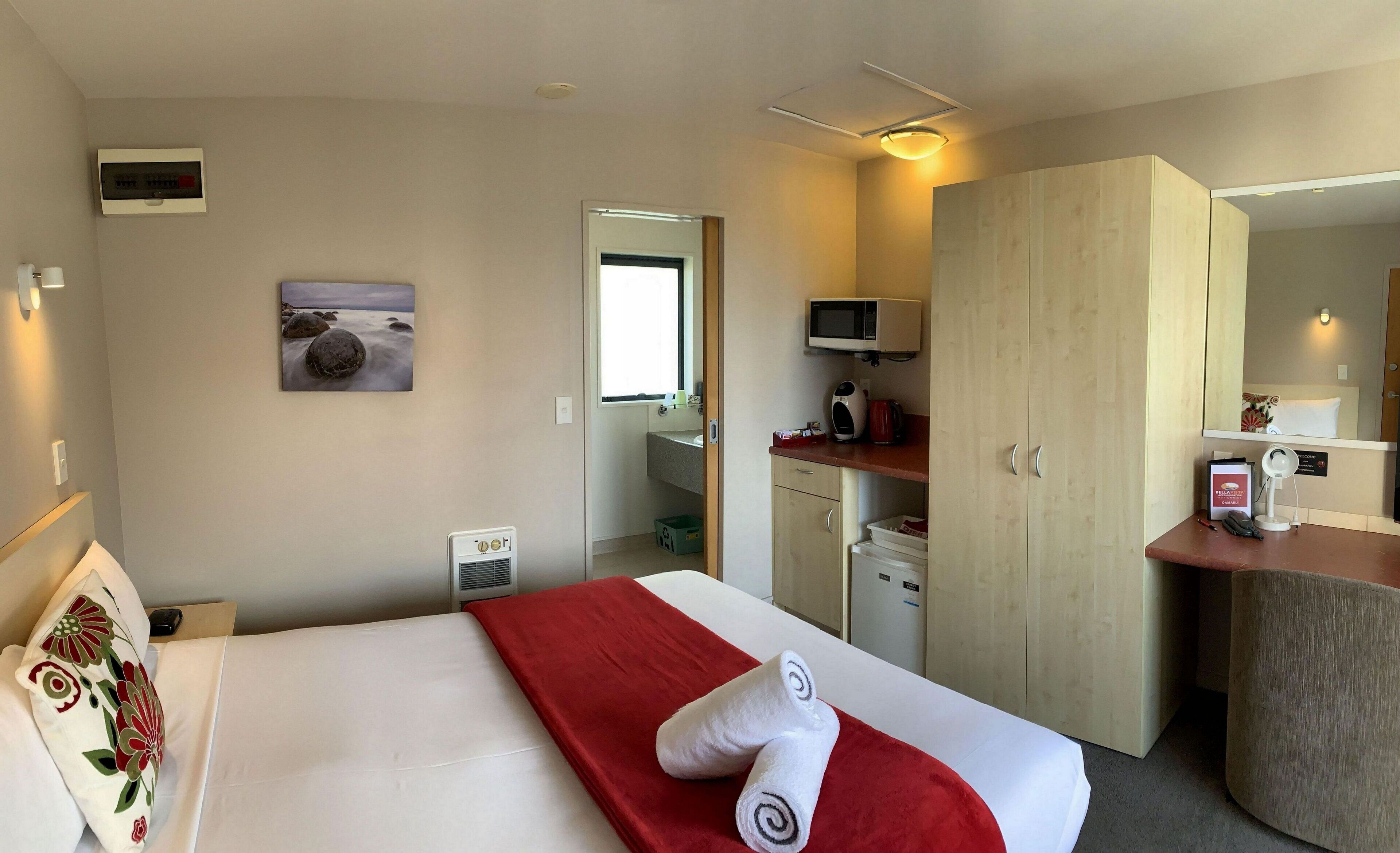 Quarto de hotel com cama, manta vermelha, televisão, minibar e banheiro visível através de uma porta aberta.