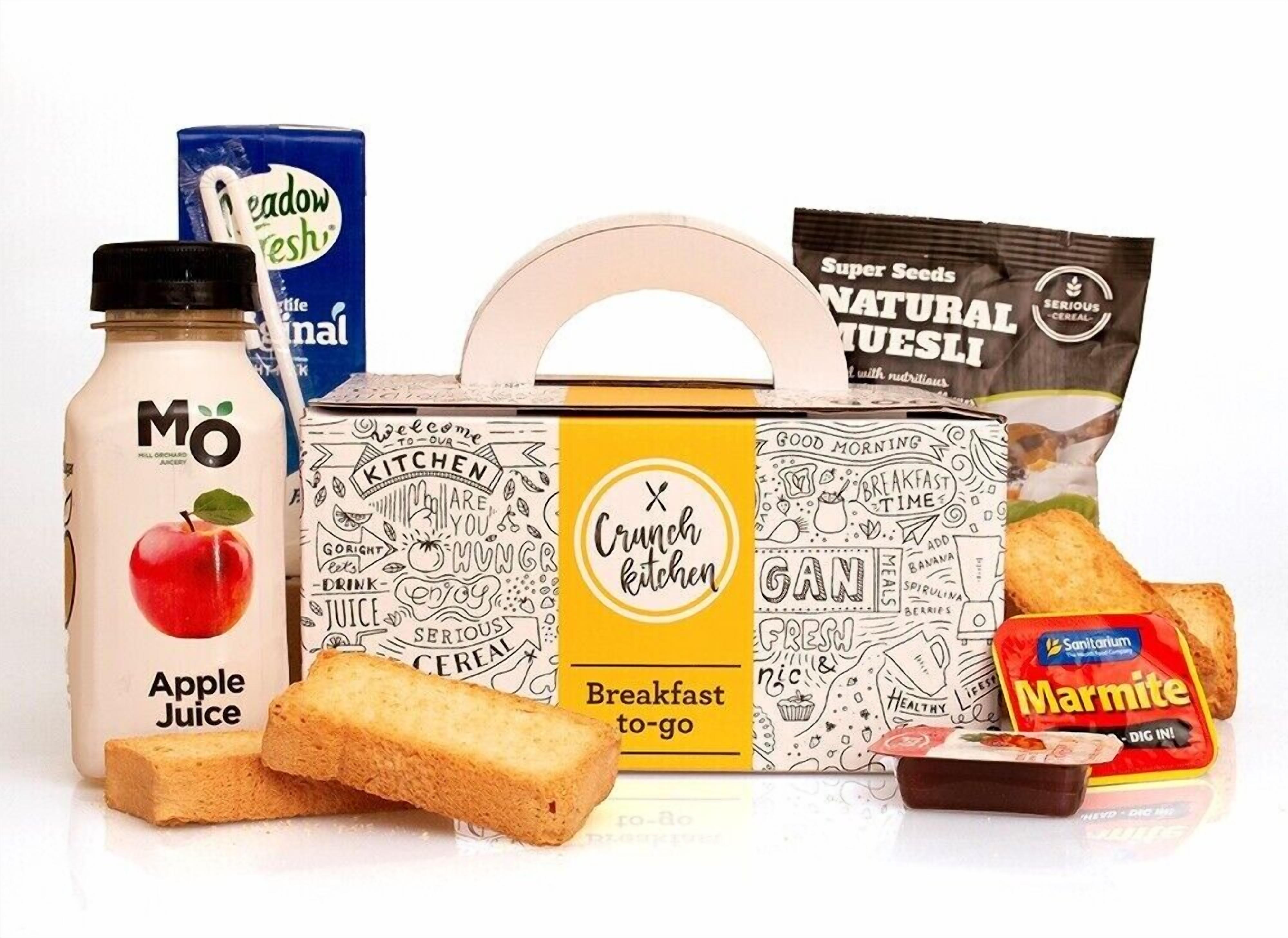 A breakfast to-go box with a handle, a bottle of apple juice, a pack of marmite, and a box of super seeds cereal.