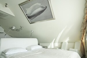 Uma cama bem arrumada com roupa de cama branca, uma mesa de cabeceira com luminária e uma obra de arte emoldurada acima.