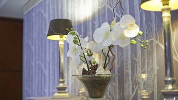 Un jarrón de vidrio con orquídeas blancas y una mariposa sobre una mesa, rodeado de lámparas y un papel tapiz estampado.