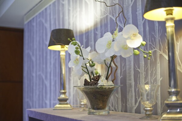 Un jarrón de vidrio con orquídeas blancas y una mariposa sobre una mesa, rodeado de lámparas y un papel tapiz estampado.