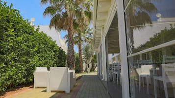 Un chemin mène à un espace extérieur aménagé avec des chaises et des tables blanches, entouré d’une végétation luxuriante et de palmiers.