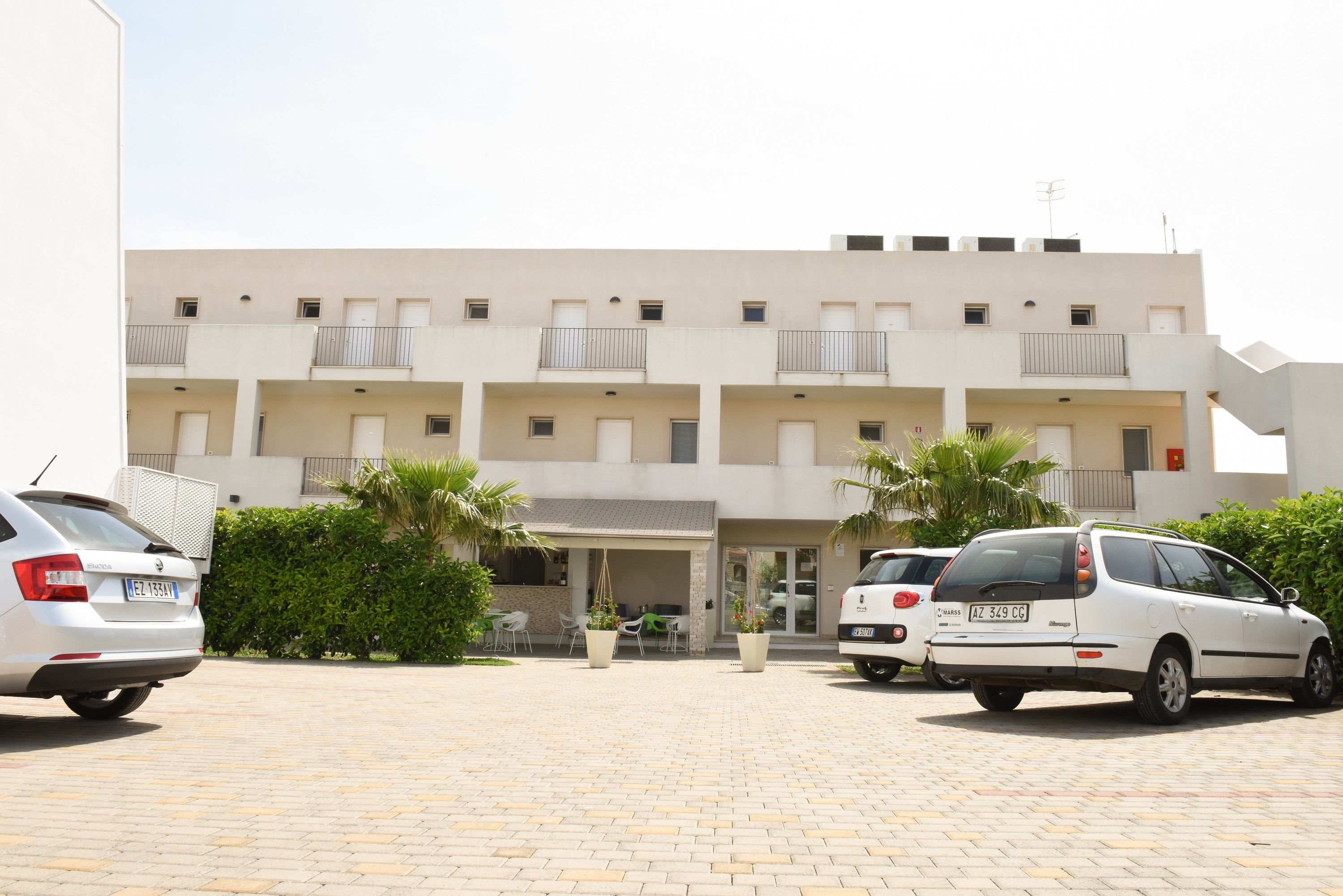 A multi-story building with a parking area in front. There are two cars parked, a white hatchback and a white station wagon. The building has balconies on each floor.
