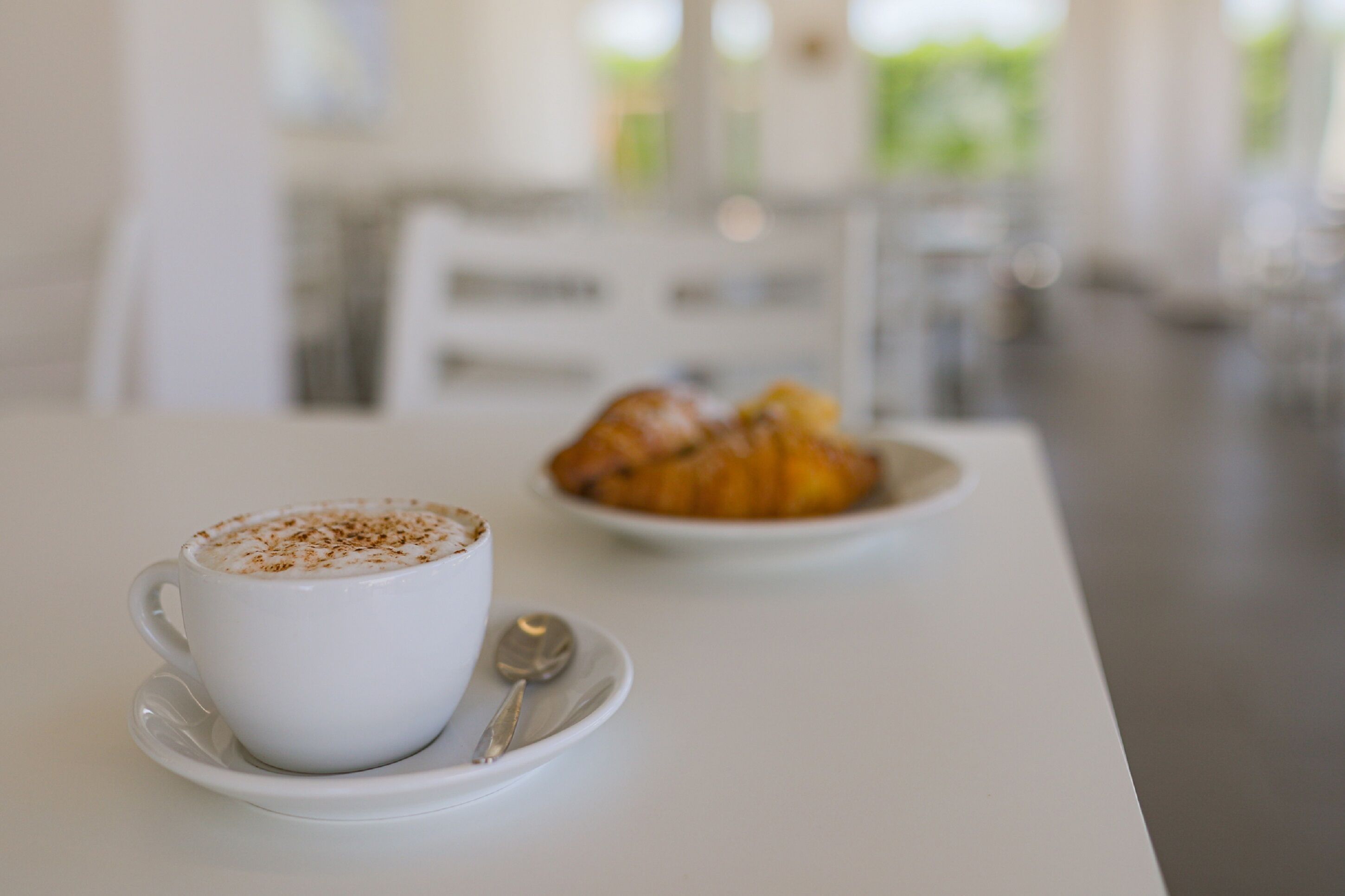 Een witte koffiekop met lepel op een schotel, en een bord met croissants op een tafel.