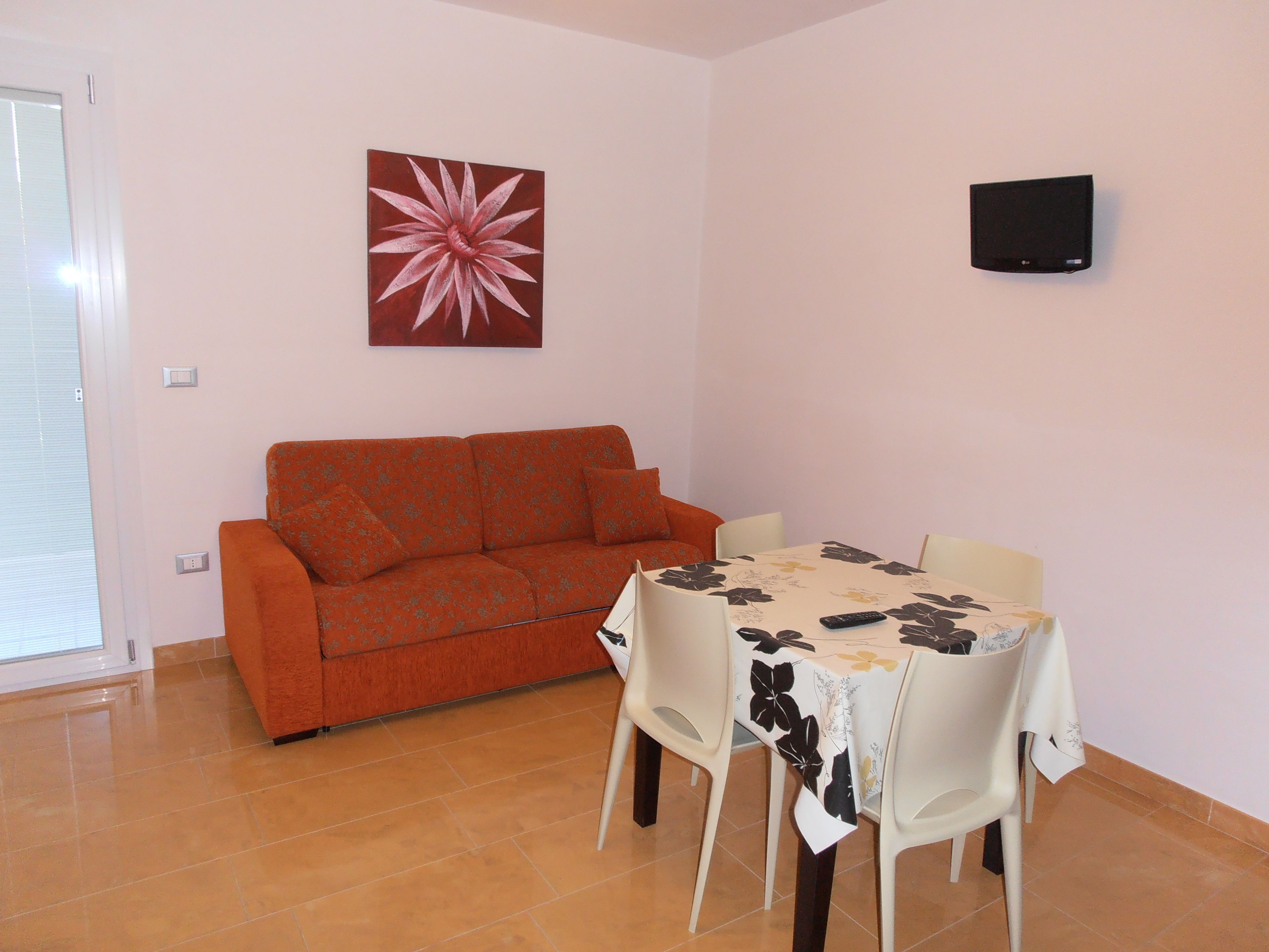 A living room with a sofa, a dining table with chairs, a wall-mounted TV, and a large flower painting.